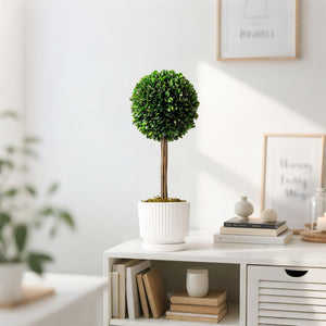 An artificial boxwood topiary tree with a round green ball on a natural wooden trunk, placed in a white ridged ceramic pot. The tree is displayed on a white console table in a brightly lit, modern living room setup with books, candles, and framed wall art in the background.