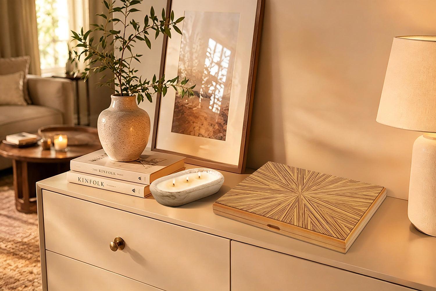 A Woodronic wooden board game box with an elegant, sunburst-patterned inlaid wood lid, displayed on a cream-colored sideboard. The warm-toned living room scene includes a speckled ceramic vase with green branches, Kinfolk books, a multi-wick candle in a stone bowl, and a table lamp, all bathed in soft, golden sunlight.