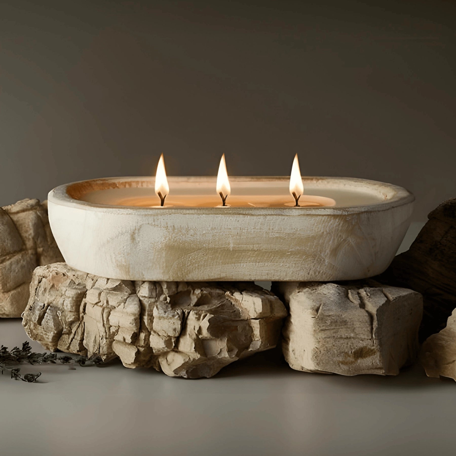 Lit three-wick candle in white-washed dough bowl, styled on natural stones for rustic home decor.